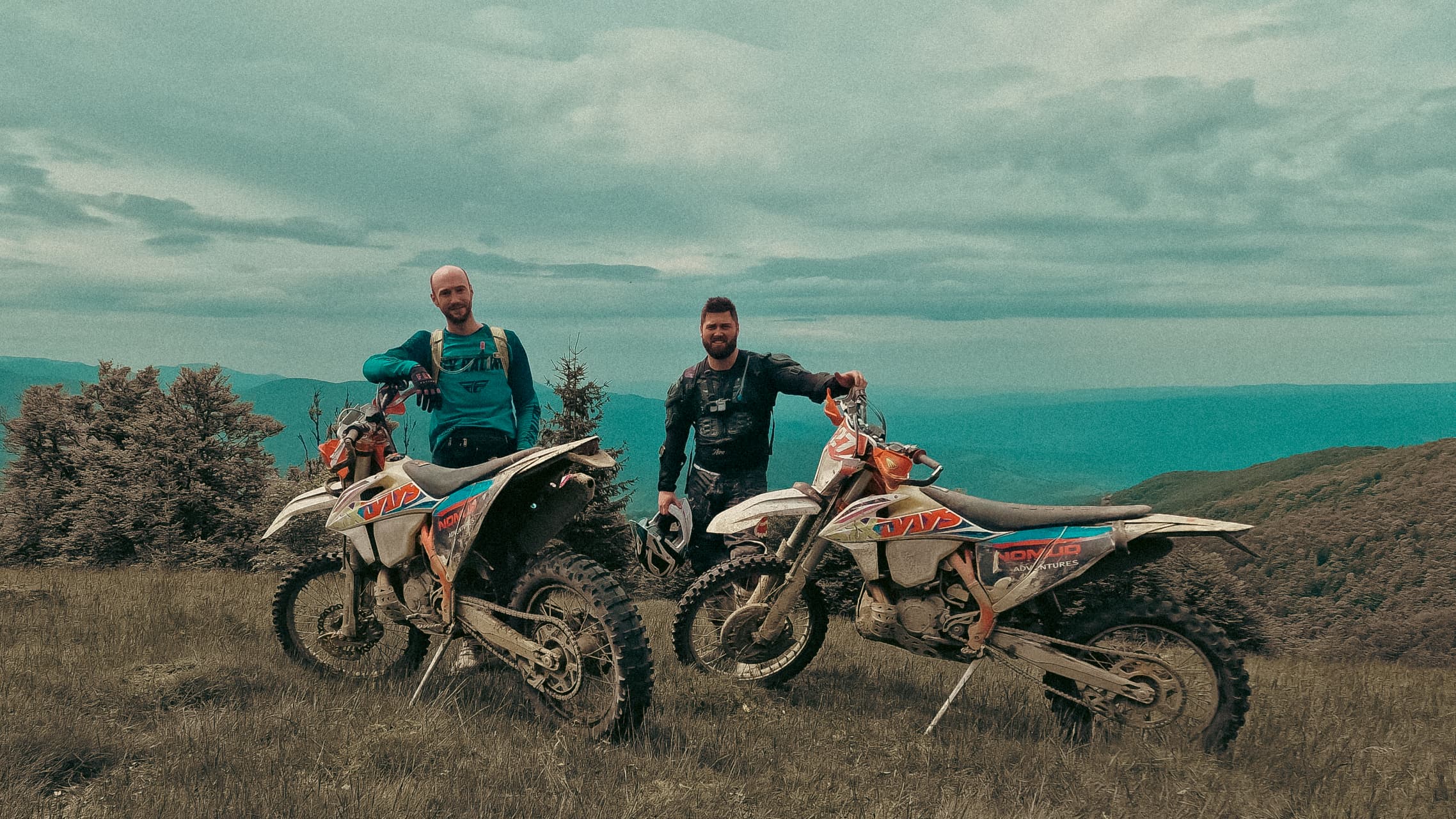 Ben and Tom standing with their KTM enduro bikes on a mountain ridge